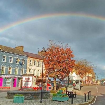 A vibrant street view of the Square in Dunmanway, West Cork, featuring a row of brightly painted colorful buildings under a prominent, full rainbow arching across a dramatic cloudy sky